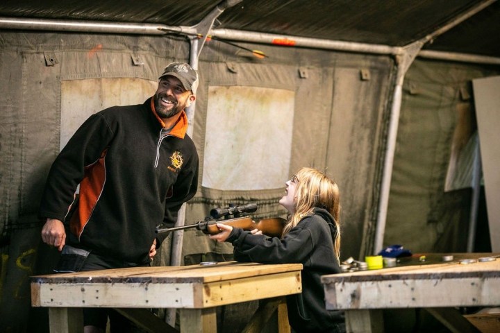 Man watches girl aiming a rifle at a shooting range.