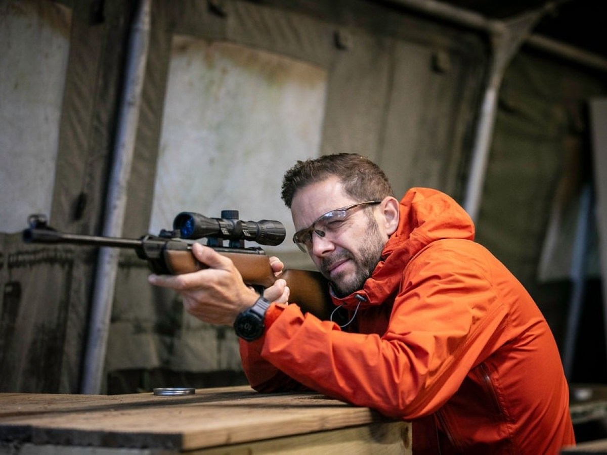 Man in orange jacket aiming a rifle with a scope, wearing safety glasses indoors.