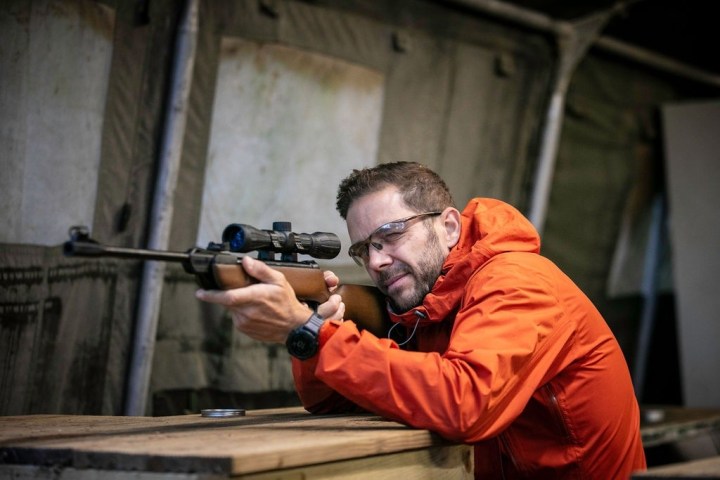Man in orange jacket aiming a rifle with a scope, wearing safety glasses indoors.