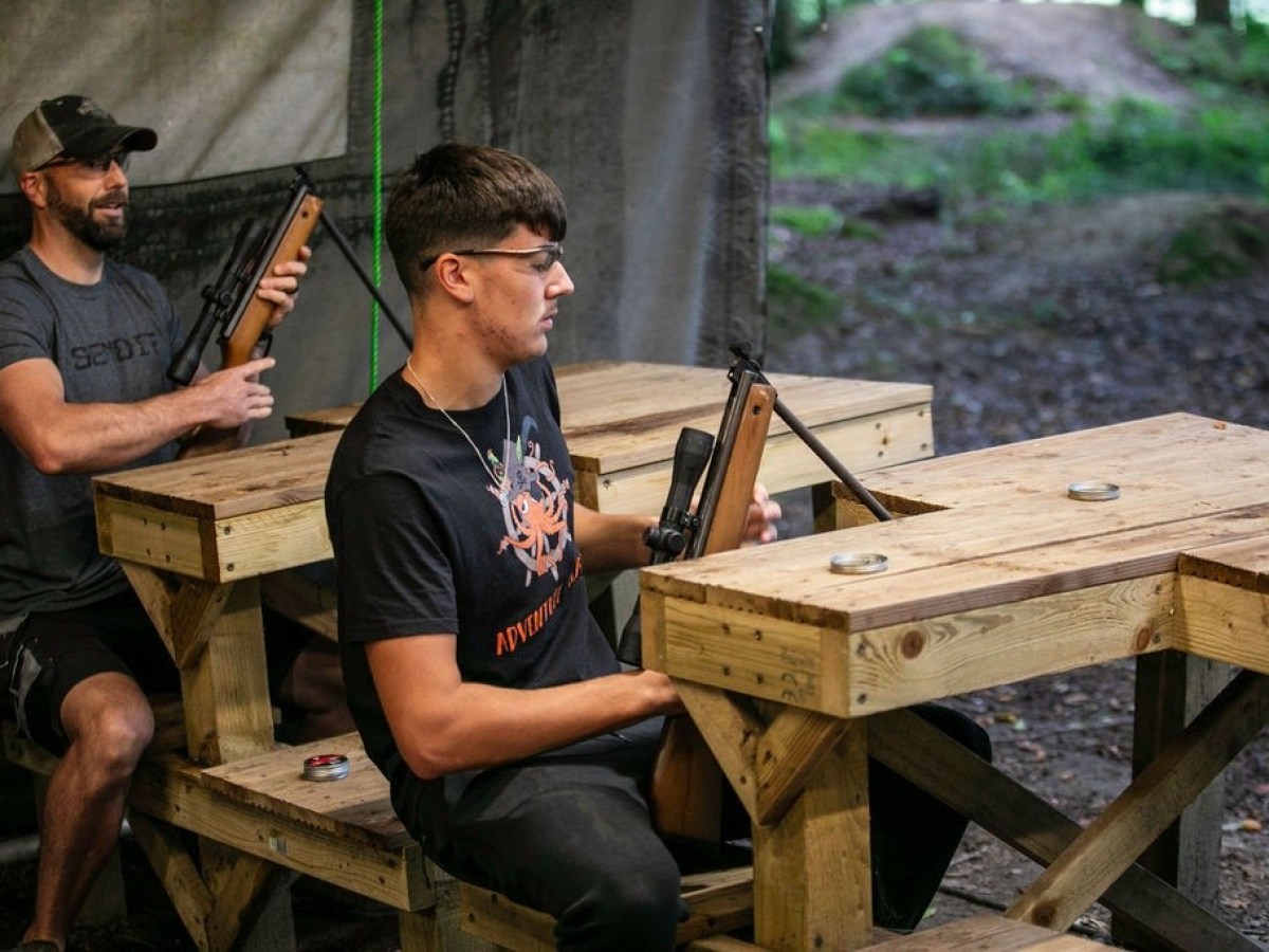 Two men seated at wooden tables aiming air rifles, outdoors.