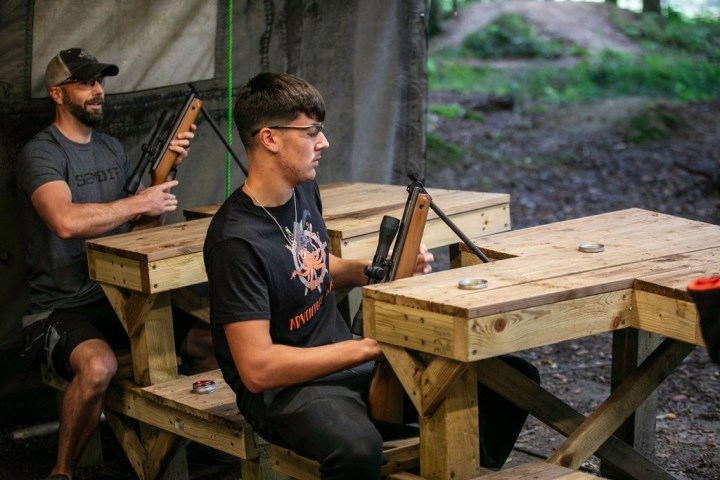 Two men seated at wooden tables aiming air rifles, outdoors.